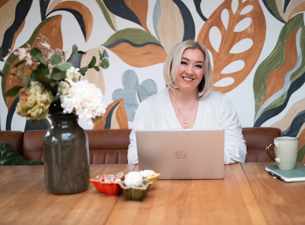 Nichola Doyle, Director of UK Therapy Guild Counselling Services, smiling at a table with a laptop in a bright room.